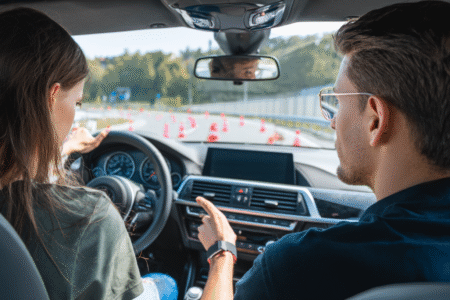 Zwei Personen sitzen im Auto, eine Person am Steuer, die andere zeigt nach vorne in Richtung Fahrbahn mit Verkehrskegeln.