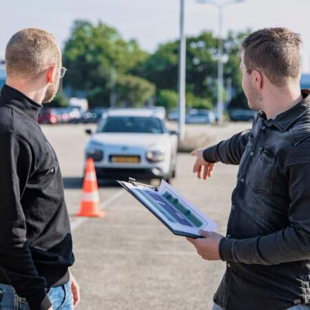 Zwei Personen stehen auf einem Parkplatz mit Verkehrskegeln, eine Person zeigt in Richtung eines Autos und hält ein Klemmbrett.